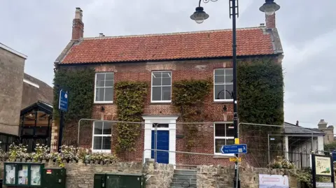 Conor Ogilvie-Davidson St Dunstan’s House which is a grand red brick building with five sash windows and a blue door with a white surround. It is partially covered in Ivy. There is some metal fencing around it preventing access.