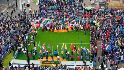 Homeless World Cup The Homeless World Cup. At the centre is a rectangular green field, bordered by fencing, where numerous national flags are displayed on poles. Inside the fenced area, people are standing in a semi-circle, each holding a flag, depicting different countries. There are crowds of people in colourful clothing in tiered stands.