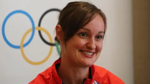Getty Images Susan Egelstaff smiling in front of the Olympic rings logo. She is wearing a bright red athletic jacket with a black zipper and a hint of a white and blue visible near the collar. 