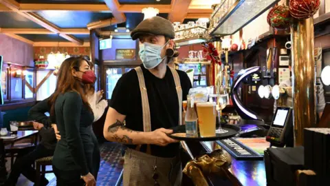 Getty Images A bartender in vintage trousers, black T-shirt and a dark green flat cap wears a face mask as he carries a tray of drinks towards customers 