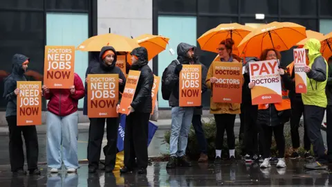 EPA Several doctors on strike, wearing orange BMA hats and hooded raincoats and holding up placards calling for more pay, saying 'patients need doctors, doctors need jobs'' outside University Hospital Liverpool on Friday.