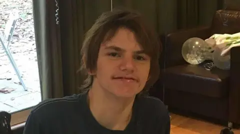 Trudy Polkinghorn A man/teen is in a room with a leather chair behind. He has dark hair and is wearing a dark top. He is smiling. There's a big glass door behind looking into a garden.