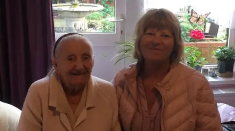Karen Staniland An elderly woman in a white cardigan and a younger woman in a coat and with lighter hair. They're in a room with a patio door behind, both sat on a bed. 