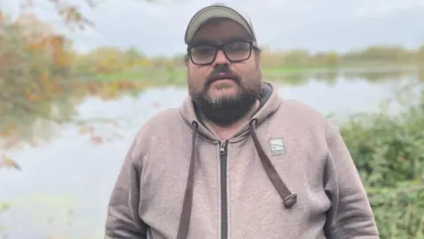Sam Read/BBC Craig Parkins, standing outside, wearing a cap, with glasses on, wearing a pale hoodie, with a beard. He is standing in front of a lake, with grass to the side. 