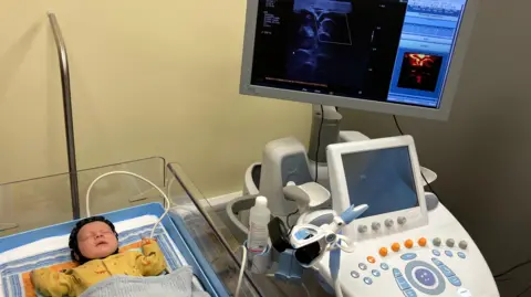 Baby Theo is lying, asleep, in a hospital cot. He is wearing the small black sensor cap, which is linked to neighbouring machines by a white cable. The monitors attached to the machines are showing scan images of Theo's brain.