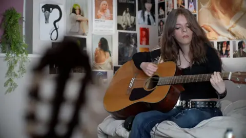JAMIE NIBLOCK/BBC Amy sits on her bed in front of a range of Billie Eilish posters on her bedroom wall. She is holding a wooden acoustic guitar and is looking at her fingers playing it. There is a green plant to the left of the photo and another guitar to the left foreground. To the right Amy sits. She is wearing blue jeans, a black and silver-coloured belt and a black top.