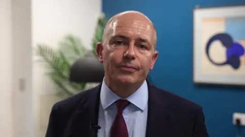 Mr Sinker is dressed in a dark suit jacket with a white shirt and a deep red tie. He is indoors in front of a blue wall. Behind him, there is a large framed abstract artwork. A tall green plant and a black floor lamp are visible to the left. His head is tilted slightly as he looks directly at the camera, not smiling, as he speaks to the BBC interviewer, who is out of shot.