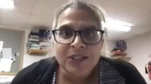 ORTHOPAEDIC ACADEMY Kuldeep Stohr looks at the camera during an NHS video she is taking part in. She is wearing a black and white blouse and a black cardigan. She has black, plastic-framed glasses on and greying hair which is tied back. She is sitting in an office with folders packed onto shelves in the background and some papers pinned to a board.