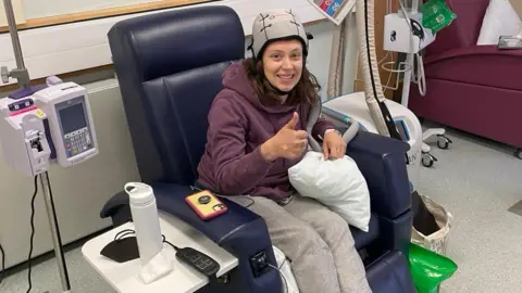 Kat Denisi Kat is in a hospital chair. She is wearing a cold cap to help with hair loss during chemotherapy treatment. She is wearing a maroon hoodie and grey trousers. There is a water bottle beside her and hospital equipment.