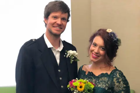 Kat Denisi Neil has short brown hair and is smiling at the camera. He is wearing a dark wedding suit and has a white flower in his button hole. His wife Kat is in a lacey green dress. She is also smiling at the camera. She has long brown hair tied up with flowers in it. She is also holding a colourful bunch of flowers.