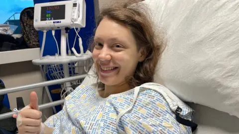 Kat Denisi Kat is in a hospital gown in bed. She has brown hair and is smiling. There is a hospital monitor in the background.