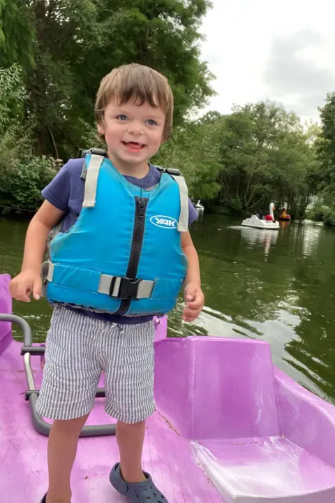 Tammy McDaid A little boy with light brown short hair is smiling while standing on a pink pedal boat on a lake. He is wearing a blue T-shirt, striped blue shorts and a turquoise life jacket. There are large pedal boats shaped like swans and other birds behind him.