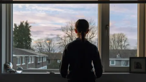 Getty Images A young person with long hair tied in a ponytail looks of her bedroom window. They are in shadow, while the roofs of other houses are clearly visible and there are trees in the background.