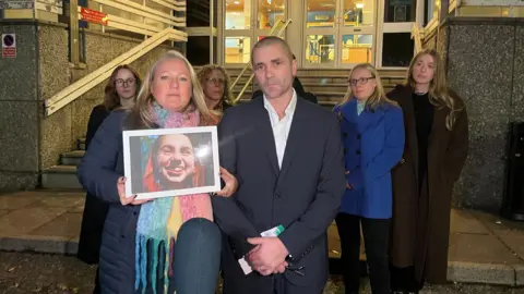 BBC / Juliette Parkin A blonde woman in a puffer jacket and colourful scarf is holding a large photo of a girl with dyed red hair. She is standing next to a man wearing a black suit and white shirt. There are four other people standing behind them. The group is standing outside a magistrates' court.