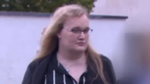 Rose Henderson arrives at the tribunal. Rose has long blonde hair, glasses and is wearing a black and white stripy dress.