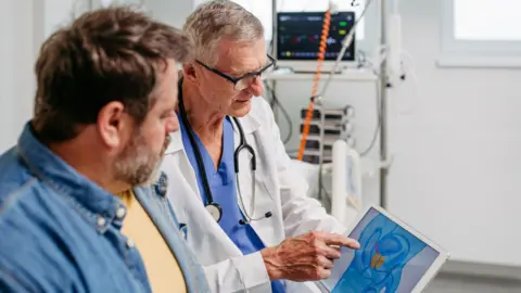 Getty Images A doctor wearing a white coat and a stethoscope around his neck looks at a tablet showing the location of the prostate, with a male patient next to him. The patient is wearing a denim shirt and has short, brown hair and a greying beard.