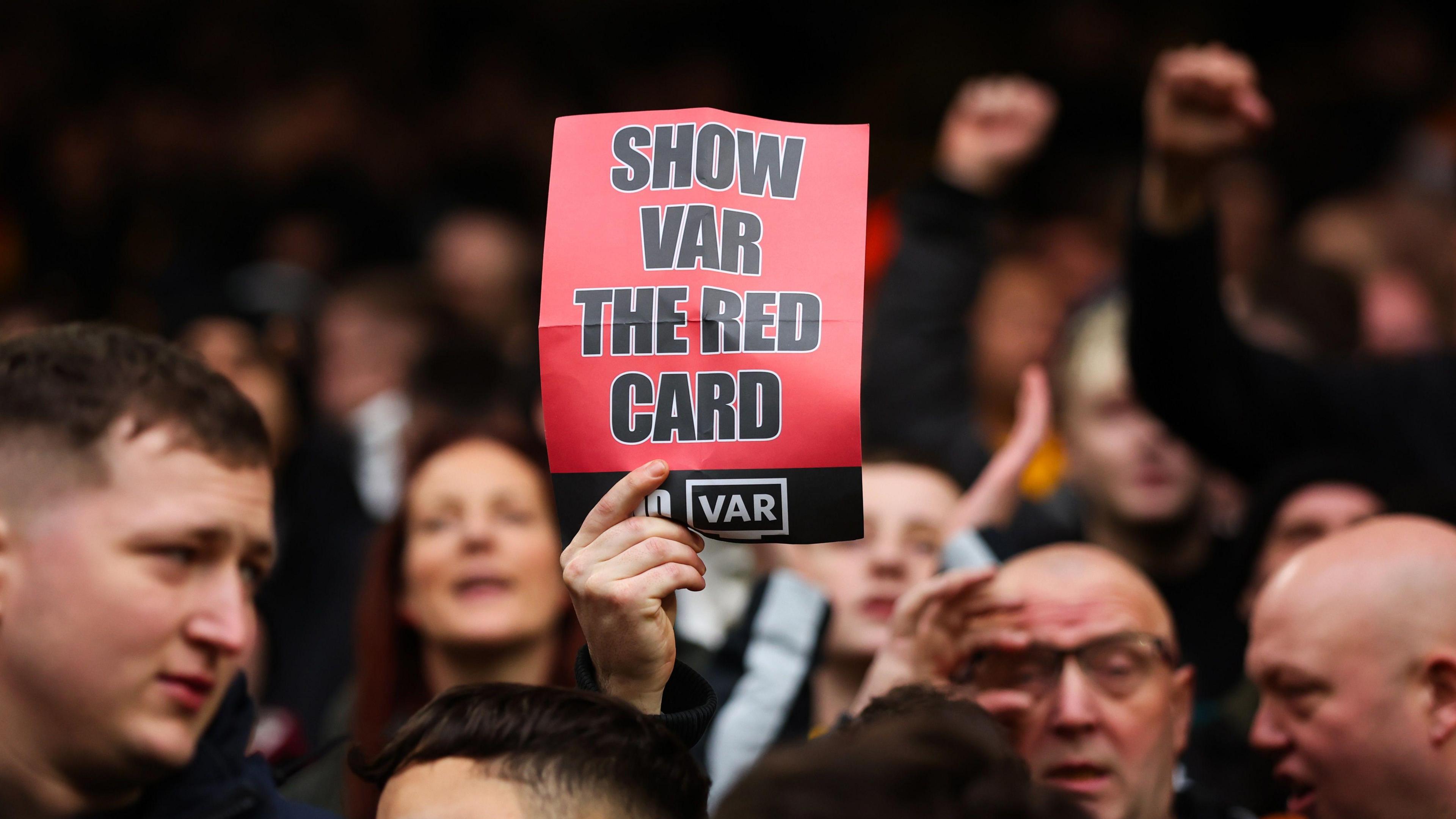 Fans hold a sign saying 'show VAR the red card'