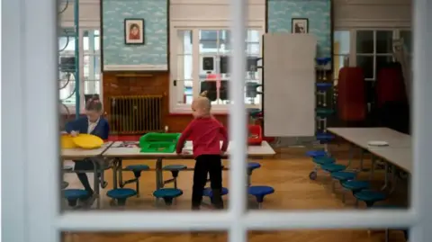 Getty Images Two children in a school classroom. The boy is wearing red and the girls is wearing blue. The camera is taking the photo from behind a window and the window can be seen in the foreground. 