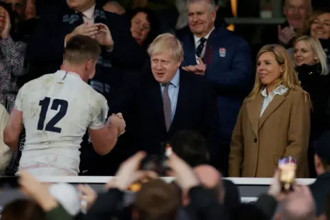 Getty Images Boris Johnson shaking hands with England captain Owen Farrell at Twickenham on March 7, 2000. Carrie Johnson, now his wife, is standing next to him.