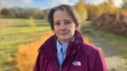 Dawn wearing a maroon North Face jacket over a light blue zip-up top, standing outdoors on a grassy path. The background features autumn foliage with golden and orange tones, scattered trees, and a slightly cloudy sky. 