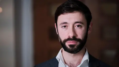 A head and shoulders photo of a man with a dark, brown hair and beard 