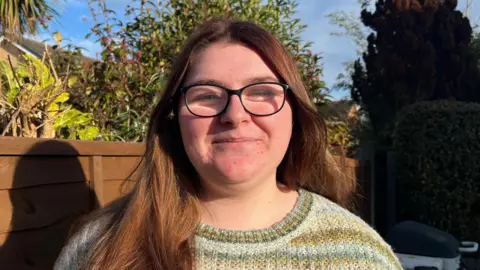 BBC/ELLEN KNIGHT Bethany pictured smiling and looking into the camera. She's wearing black-framed glasses and a striped woollen jumper in green, yellow, and white. She's stood in front of a brown garden fence with trees and shrubbery rising up behind it. The sky is blue and there's strong wintery sunshine. 