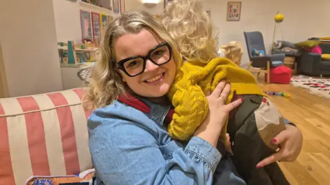 Rose Stokes A blonde woman with black, square glasses holding a blonde male toddler over her shoulder