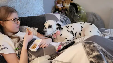 Cancer Research UK handout Sophie Claxton with long brown hair lying on the sofa under a quilt eating bread sticks and soft cheese snack while her white and black spotted dalmatian dog, Bonnie, lies next to her staring at the snack. Bonnie is wearing a pink white and black bow tie collar.