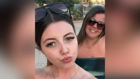 Cancer Research UK handout Sophie Claxton with long brown hair and sunglasses resting on her head takes a selfie with smiling mother Michelle Bebb with long dark brown hair and wearing sunglasses and a dark green sleeveless dress in the background.