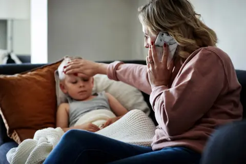 Getty Images Woman and child o a sofa. The woman is on the phone while touching the forehead of the child, who looks ill