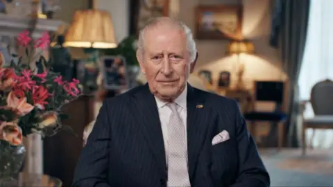 Bango Studios/PA King Charles III in The Morning Room at Clarence House, London, recording a personal message. He is wearing a dark blue striped suit and pale tie, and is sitting with pink flowers on his right and there are chairs and lamps in the background. 