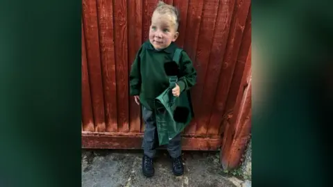 Contributed Edward is wearing a green school uniform and standing against a wooden fence.