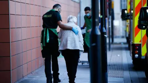 Getty Images An elderly women being led passed an ambulance by a paramedic wearing a green uniform and an apron. He has his arm around the lady's back. 