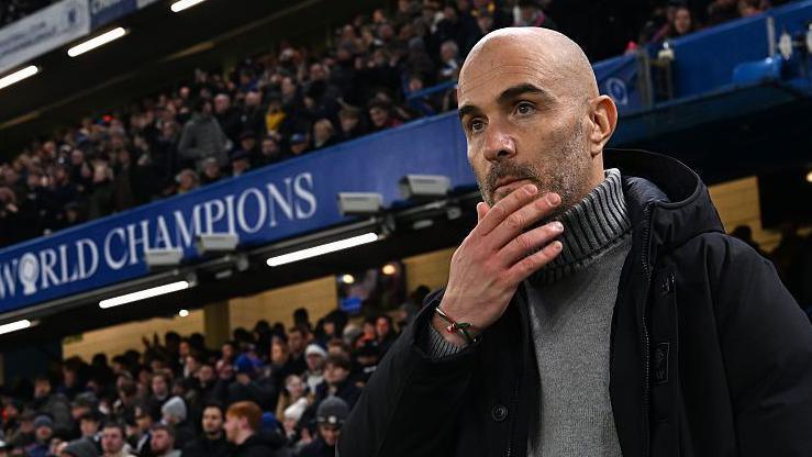 Enzo Maresca, manager of Chelsea, looks on prior to the Premier League match between Chelsea and Bournemouth at Stamford Bridge. He is wearing a grey jumper beneath a dark coat.