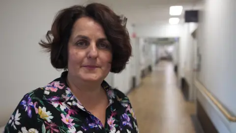 A woman with short brown hair a flowery shirt stands in a hospital corridor and looks at the camera.