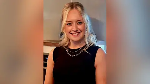 Family handout A young woman is wearing a sleeveless black top and a necklace, and has long blonde hair. She is smiling at the camera indoors.