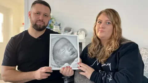 Shane Bevan and Laura Tongue hold up a black and white photo of their baby boy Louie, it is in a white frame. He is wearing a knitted hat and the photo is a close up of his face. Shane has short dark hair, brown eyes and is wearing a black t shirt and Laura has long blonde wavy hair, blue eyes and wears a navy jumper with mummy printed across it