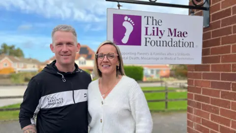 Ryan and Amy Jackson are smiling at the camera, standing next to a building with a sign reading: "The Lily Mae Foundation - supporting parents and families after Stillbirths... Miscarriage or Medical Termination". In the background are homes and a lawn