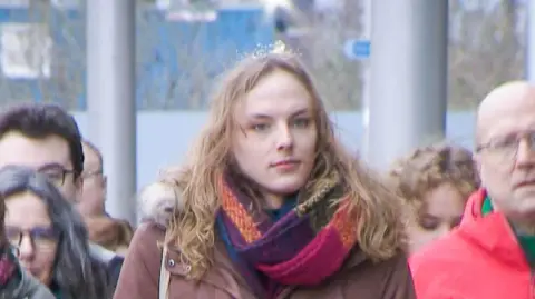 Dr Beth Upton - a trans woman with long blond hair, a maroon coat and a multi coloured scarf, walks outside along with supporters and family members.