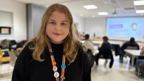 Molly has long, wavy blonde hair and is wearing a black jumper and rainbow lanyard with pin badges. She is smiling at the camera.
She is standing in a large room where other youth board members are sat, looking at a white board.
