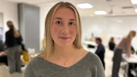 Elen has long, blonde hair and is wearing a grey jumper. She is standing in a large room where the new resources were launched, and behind her, out of focus, are the people who attended the event talking to each other at tables.
