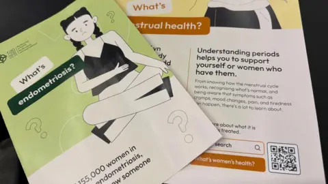 A leaflet and poster are laid out on a desk. On the top is a leaflet with the NHS Wales logo, and an image of a frowning young girl holding her hand over her tummy. It reads: what's endometriosis.
Beneath that is a poster with a QR code and a search term: what's women's health?
The poster reads: Understanding periods helps you to support yourself or women who have them.