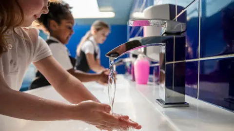 Getty Images Three children wearing school uniform wash their hands in a bathroom sink. You can't see the face of the child closest to the camera, but she has long brown hair and is running her hands under a tap, which is in focus. Behind her, another girl wearing a white polo top and a grey pinafore, with curly dark hair tied back, washes her hands. At the back, nearest the cubicles, another girl with long blonde hair - wearing the same clothes - does the same. The cubicle doors behind them are blue, as are the tiles behind the white sink. 