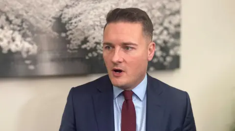 Wes Streeting speaking at a meeting. He has brown hair and blue eyes. He is wearing a navy suit, blue shirt and plum-coloured tie.