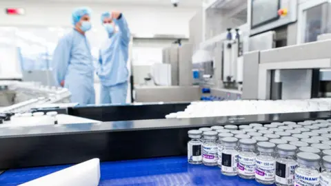 AFP via Getty Images two people in blue medical suits, hairnets and masks stand behind a conveyor belt that is covered with vials of clear medicines