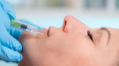 Getty Images A close-up of a woman's face, side on, getting an injection of filler into the skin surrounding her lips. The needle hovers above her lip, held by a hand wearing blue gloves. 