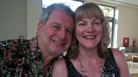 Family photo Chris and Ruth Stone-Houghton. Their heads are next to one another as they lean in for the photo. Chris is wearing a patterned shirt, and Ruth a red and white top. She is wearing a necklace and holding a wine glass.