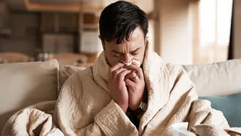 Getty Images Sick looking man wrapped up in a cream coloured blanket and holding a tissue to his nose. 