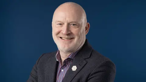 Allan Pacey Man with white stubble on his chin smiles at the camera. He's wearing a shirt and tweed jacket. The background is a blue photographic screen 