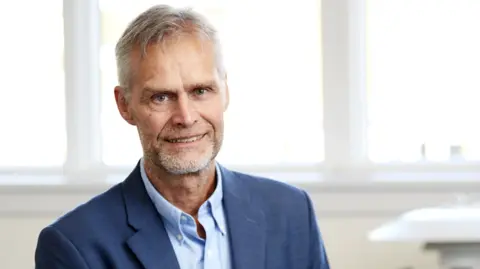 Cryos International A grey-haired man in a blue shirt and business suit jacket sits smiling at the camera with a clinical setting and big windows blurred in the background